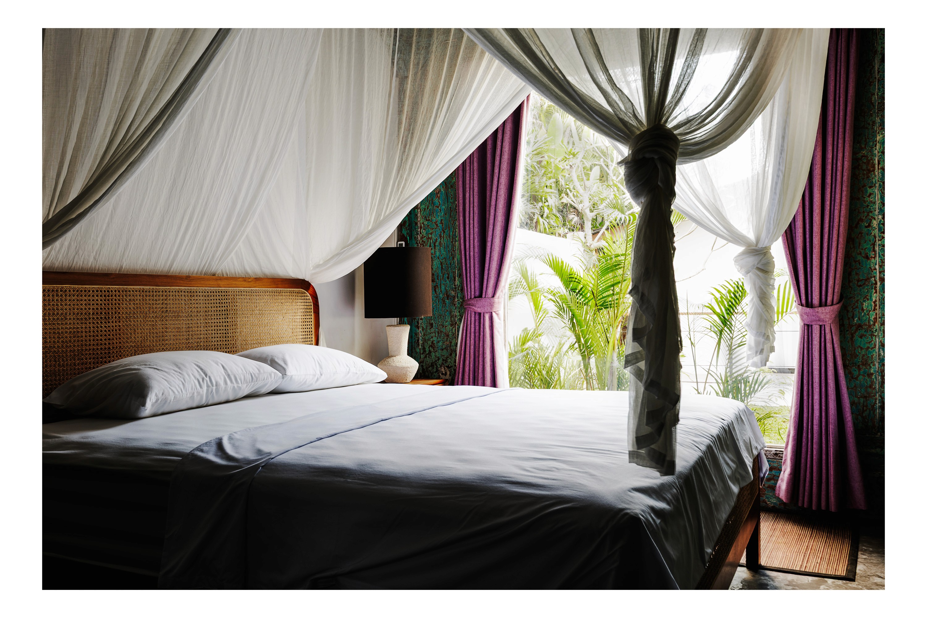 Joglo-style bedroom with rattan headboard, sheer canopy drapes, and tropical garden view through large open window.
