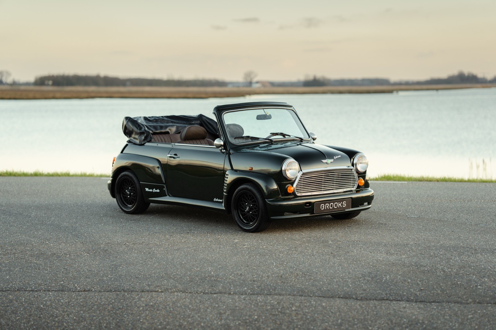 A mini classic cabrioni convertible car drives along a scenic road, surrounded by open fields and greenery.