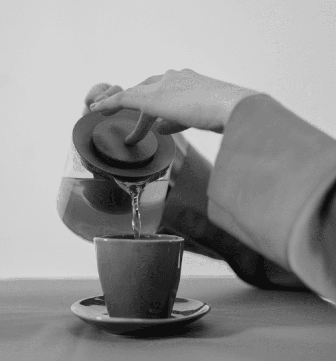 A close-up shot of a person's hands pouring liquid from a glass teapot with a round lid into a small cup on a saucer.