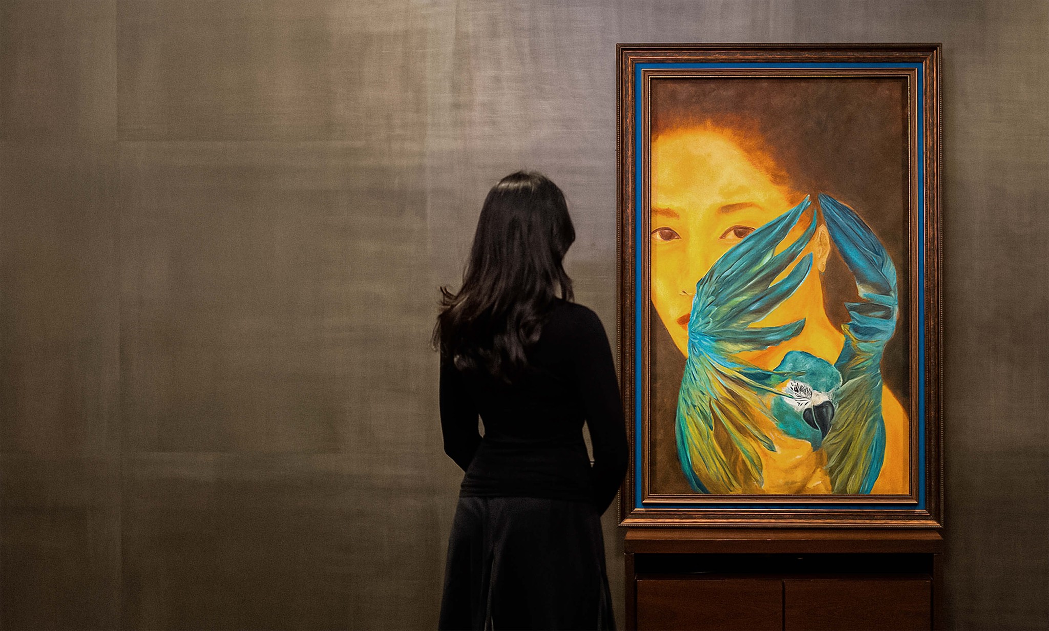 Person viewing a framed painting of a woman’s face with blue bird wings and a parrot in an art gallery.