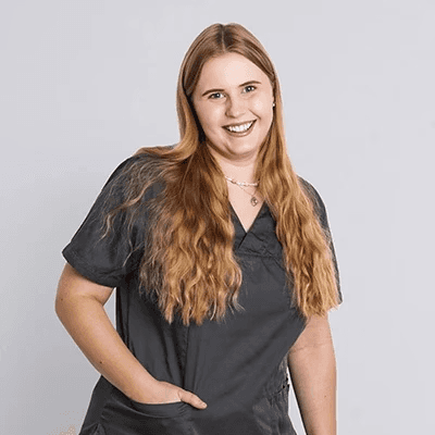 Junge Frau mit langen blonden Haaren trägt graue Arbeitskleidung und weiße Hose.