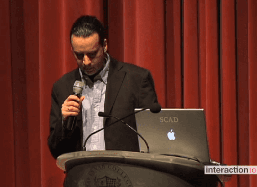 A man wearing a suit and holding a microphone giving a lecture from a laptop.