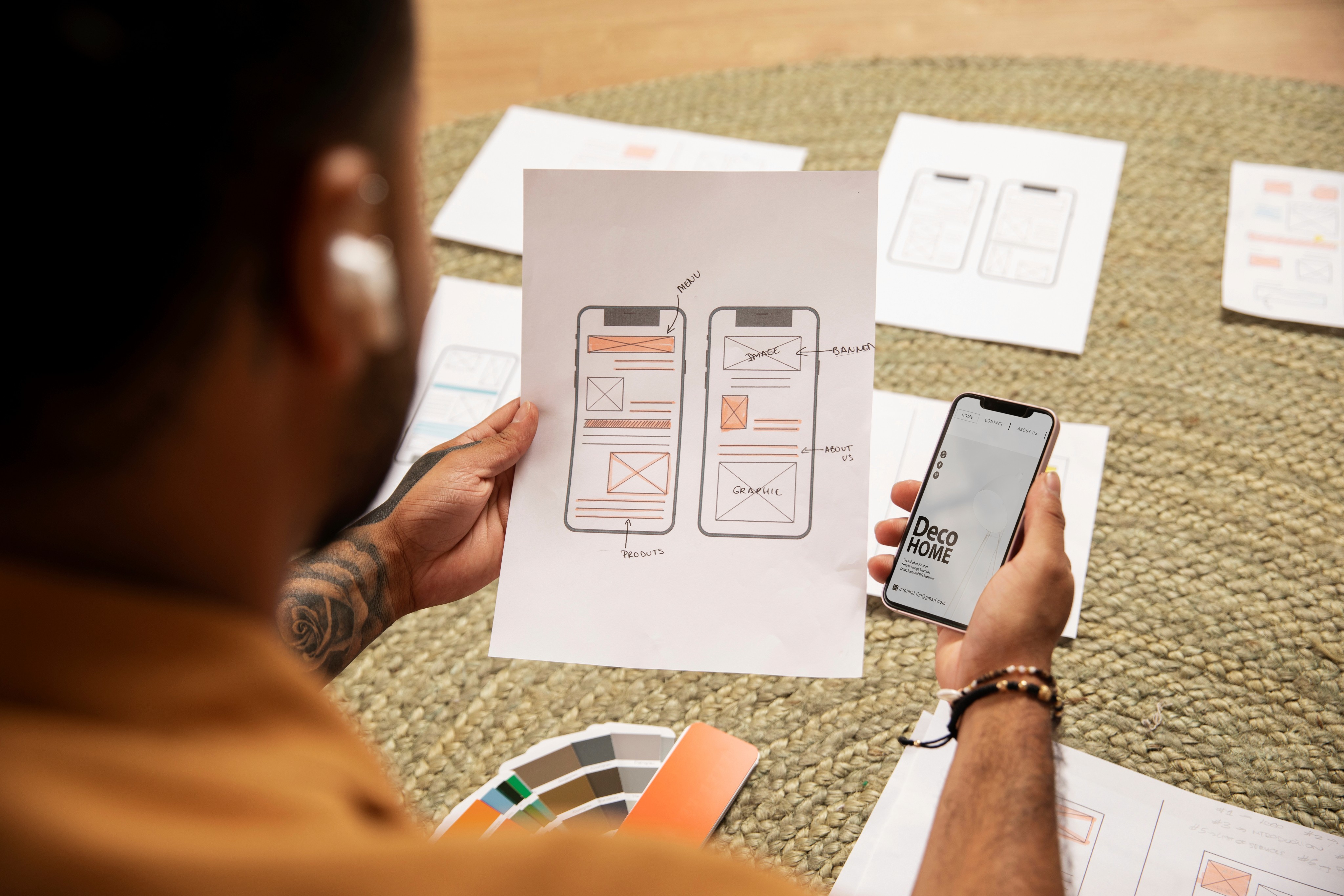 Person holding a paper with hand-drawn mobile app wireframes and a smartphone displaying a Deco Home app interface, surrounded by design sketches and color swatches on a woven surface.