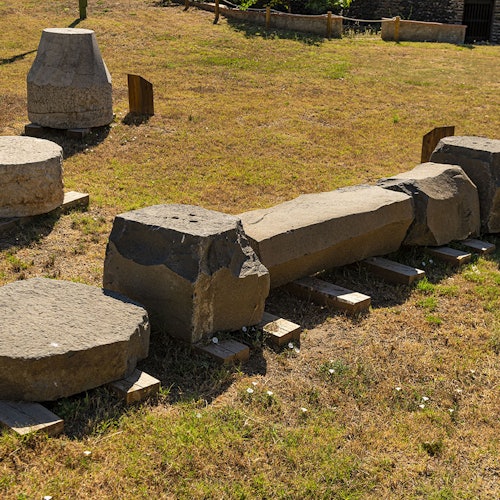 Каменные руины, включая колонны и цилиндрические блоки, разбросаны по травянистой местности.
