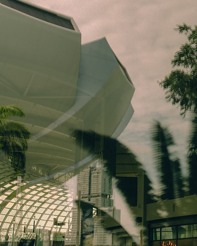 Reflection of a modern building with a unique architectural design, surrounded by greenery and a cloudy sky.