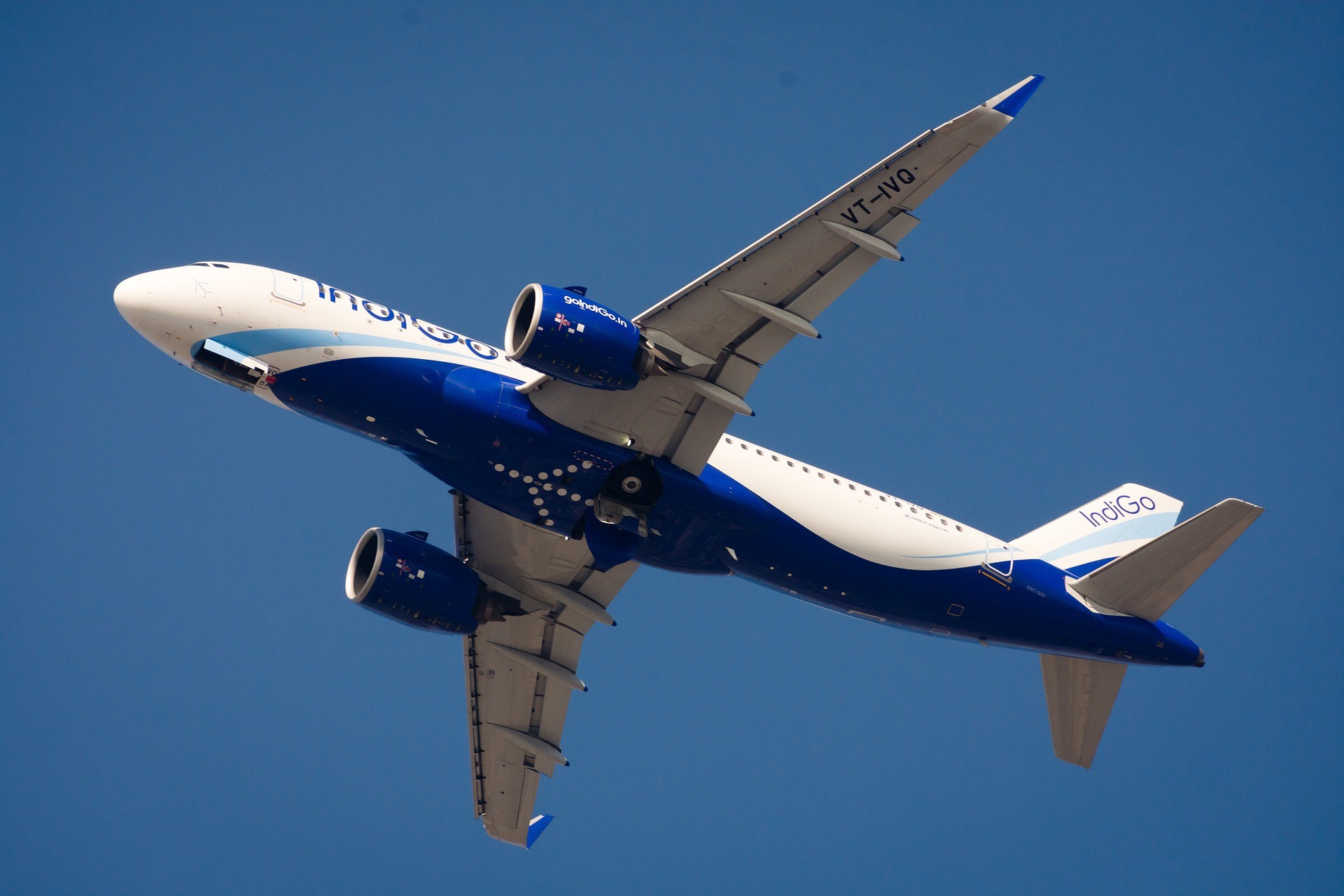 A low-angle shot of an IndiGo airlines Airbus A320 flying through a clear blue sky. The plane's underbelly is visible, showcasing its signature white and navy blue livery and the "IndiGo" logo on the tail.