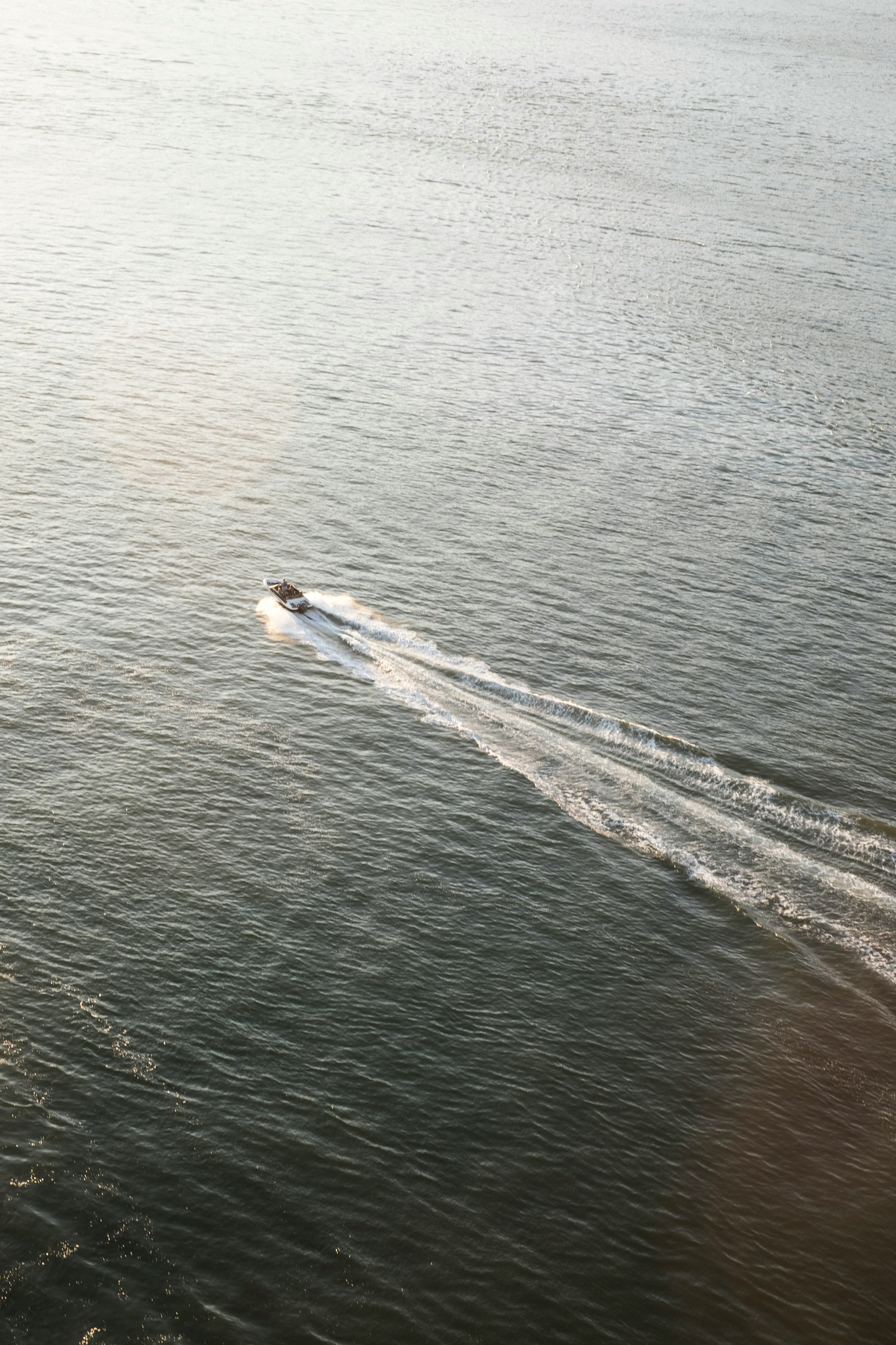 A small motorboat speeds across a vast expanse of calm, shimmering water under soft, diffused sunlight, leaving a gentle wake in its path.