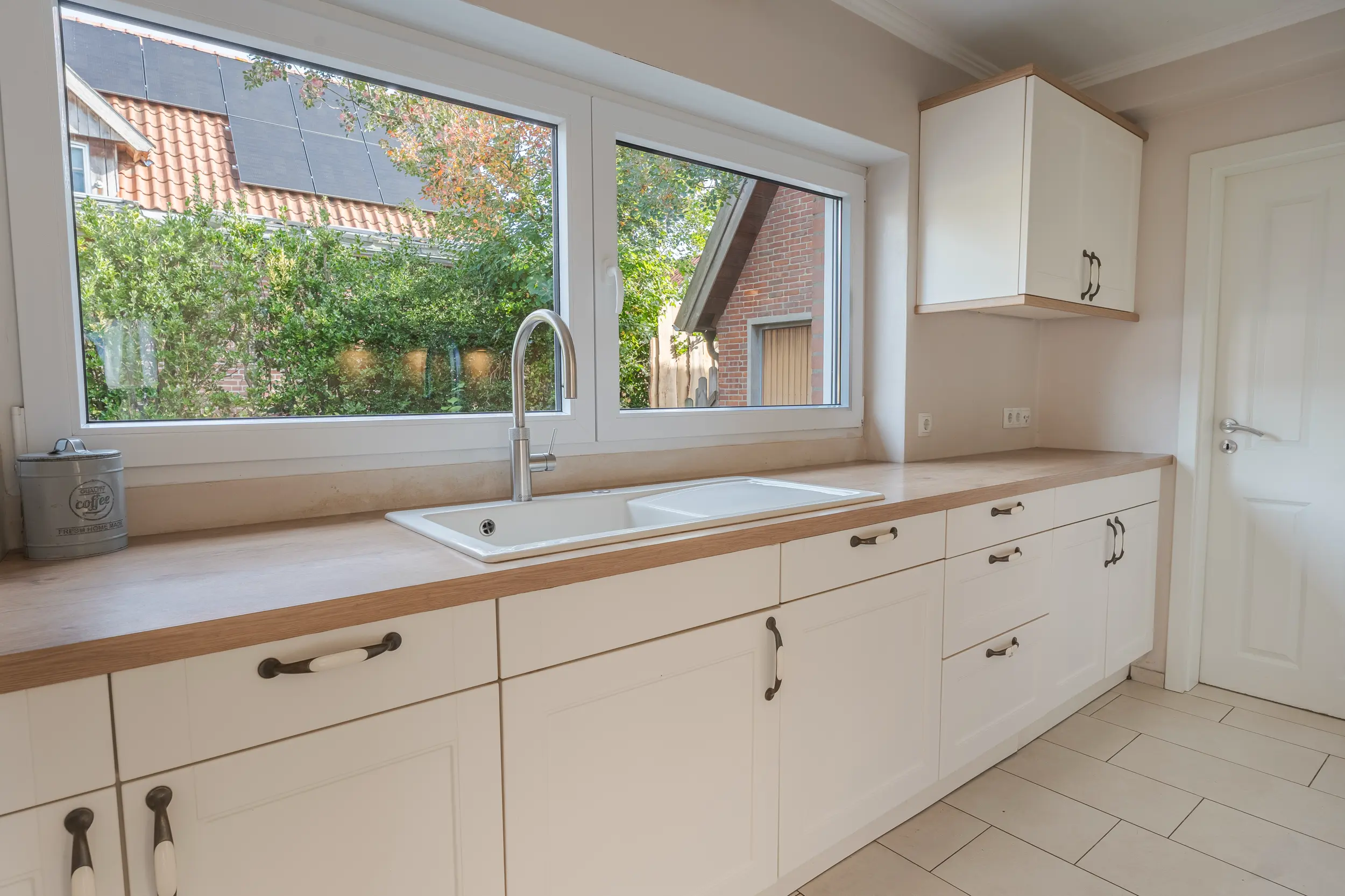 Küche im Einfamilienhaus in Südlohn-Oeding mit weißen Fronten, Arbeitsplatte in Holzoptik und Spüle vor dem Fenster.