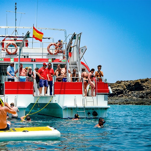身穿泳装的人，有些人在一艘挂着西班牙国旗的船上，其他人在清澈的蓝色海水中游泳，靠近岩石海岸。