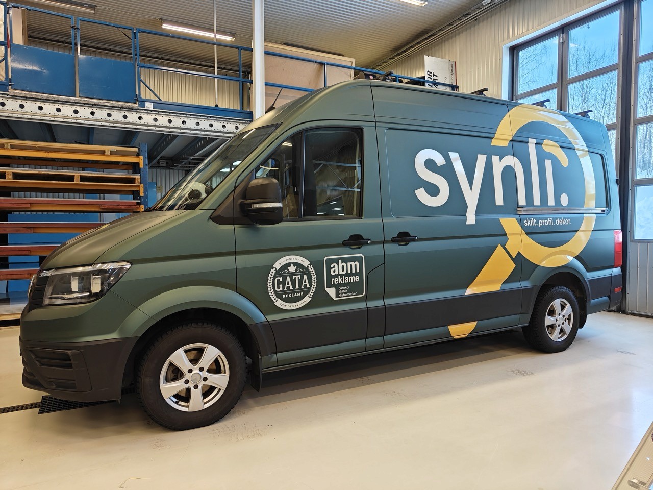 En grønn leveringsbil med merkevare for “synli” er parkert innendørs, med logoene for “GATA” og “abm reklame” på siden, mot en bakgrunn av metallhyller og store vinduer.