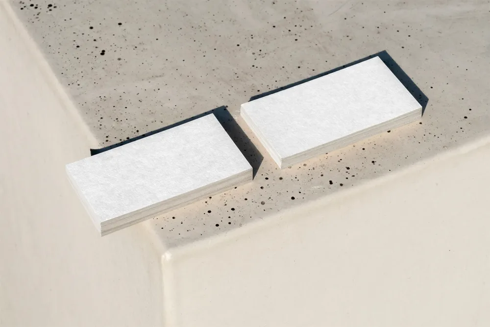 Stacked business card mockup displayed on a speckled concrete surface. One stack shows a minimalist black-and-white design on the front, while the other reveals the black back with white text. Natural lighting creates soft shadows.