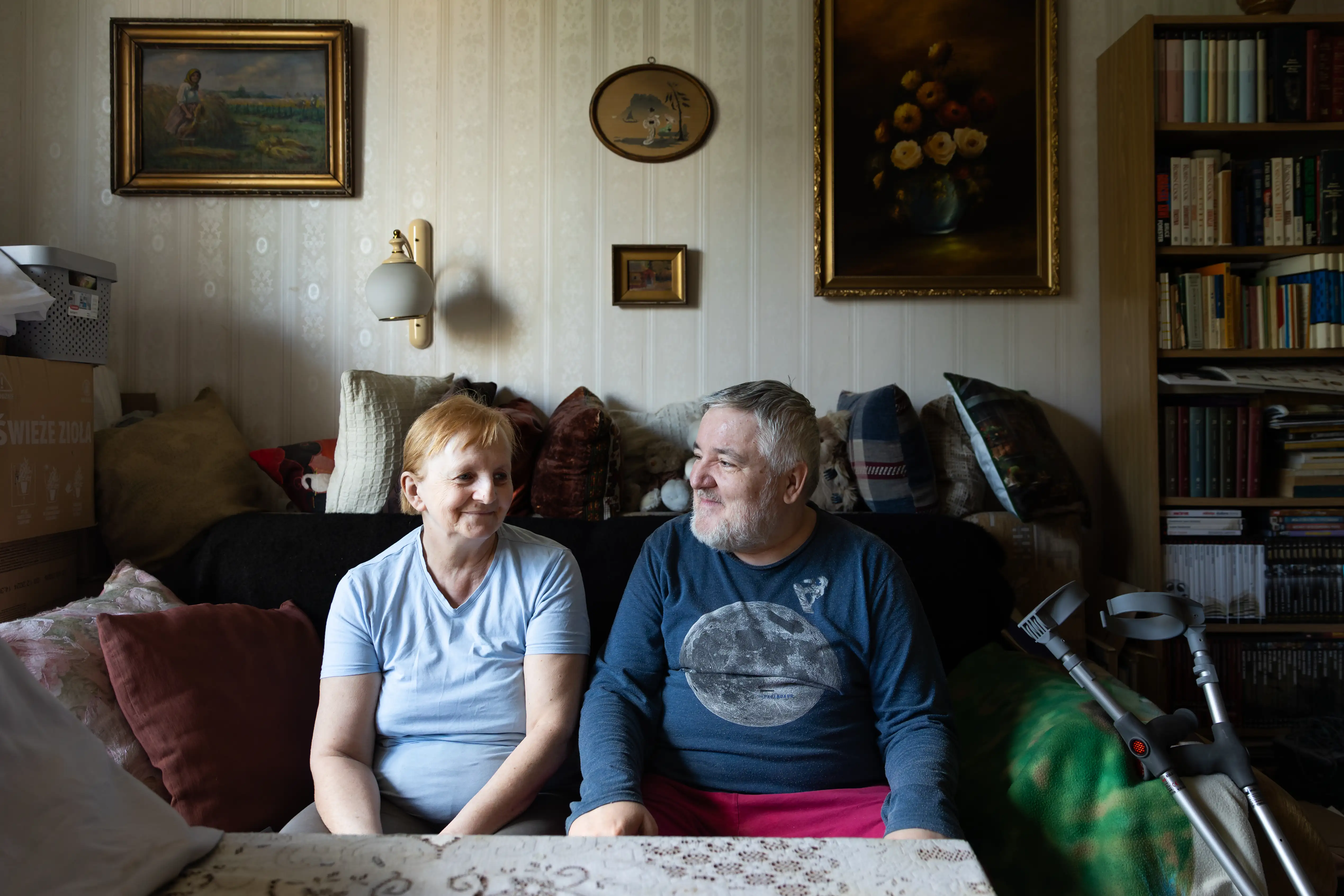 Mężczyzna i kobieta siedzą obok siebie w salonie, spoglądając sobie w oczy. W tle widać obrazy na ścianie oraz meble.