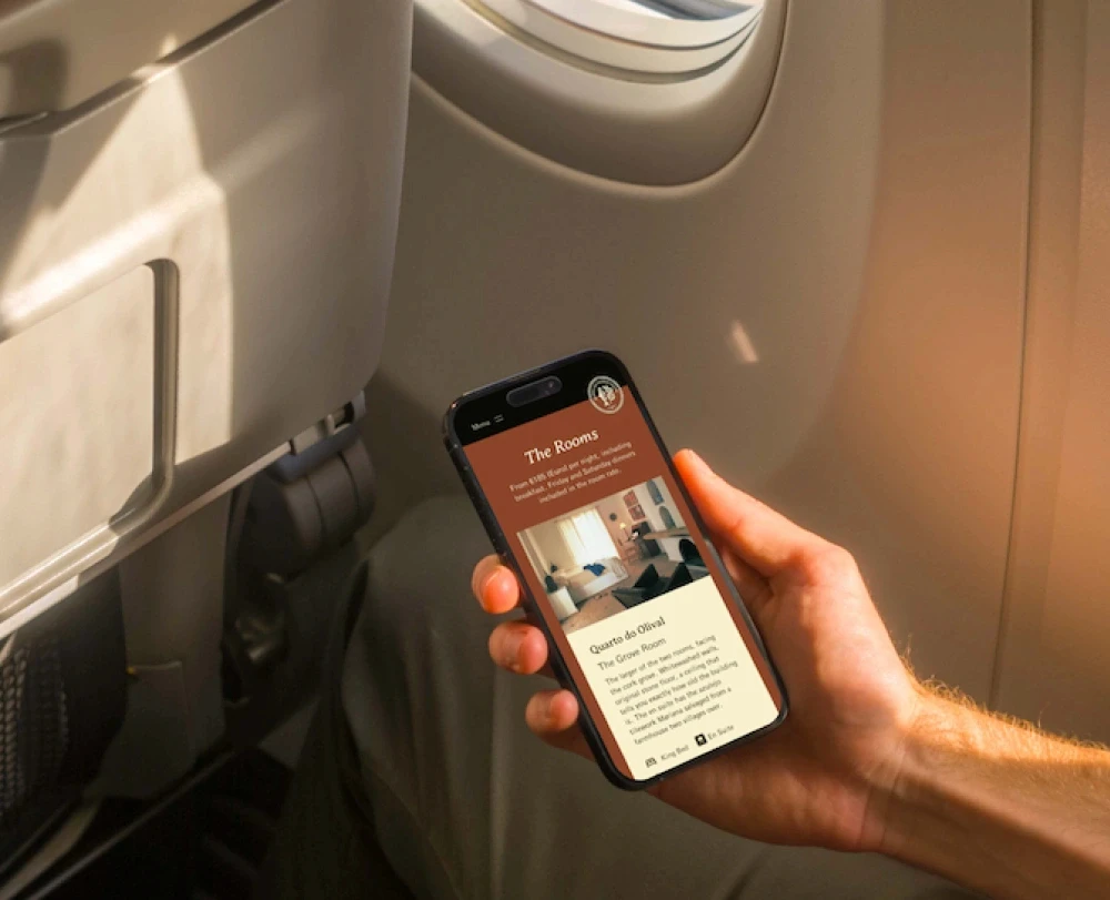 A person in a sunlit airplane seat viewing a room info card on a mobile phone.