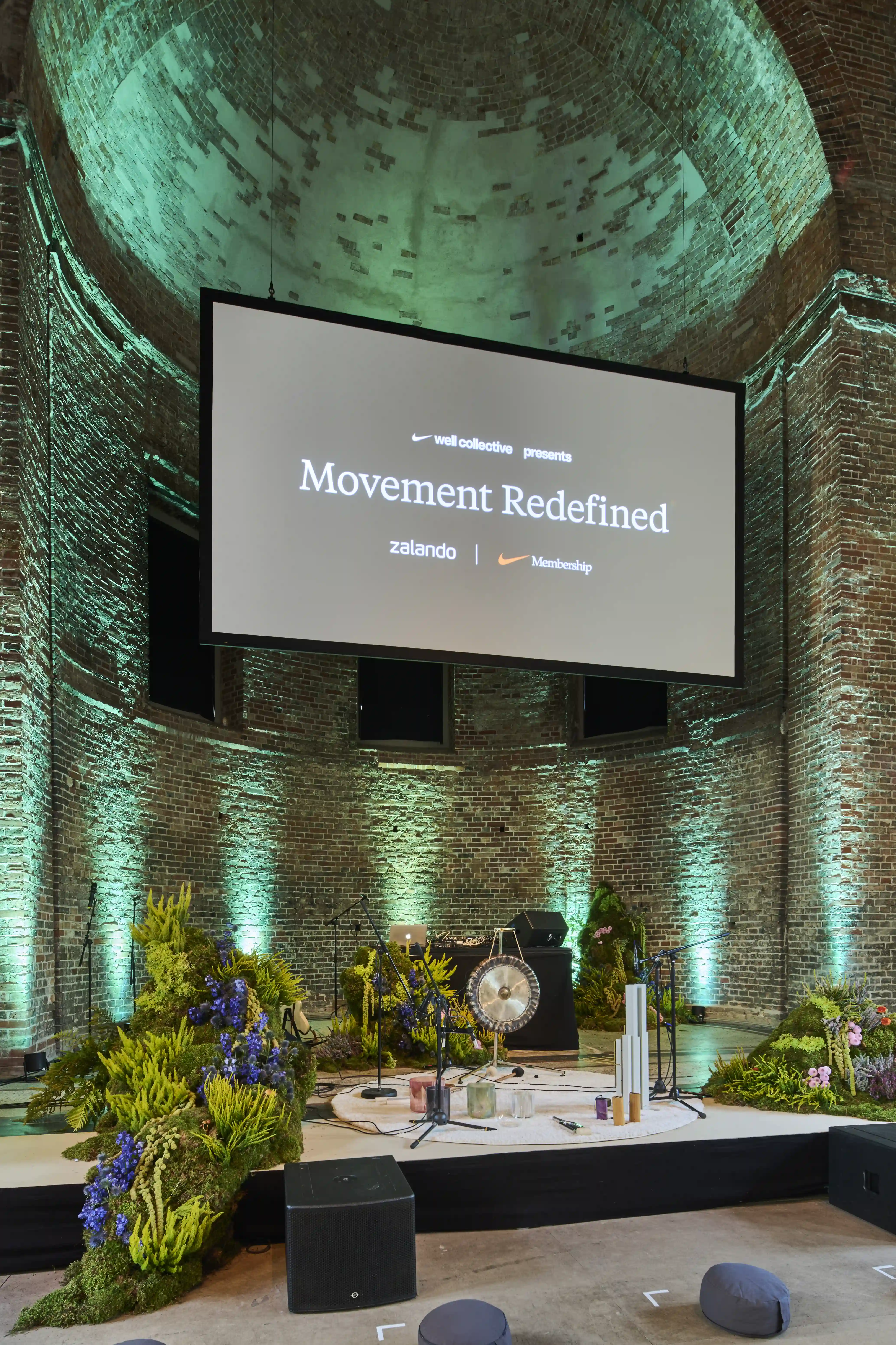 Stage in a brick rotunda with Movement Redefined screen, gong, plants and green uplighting.