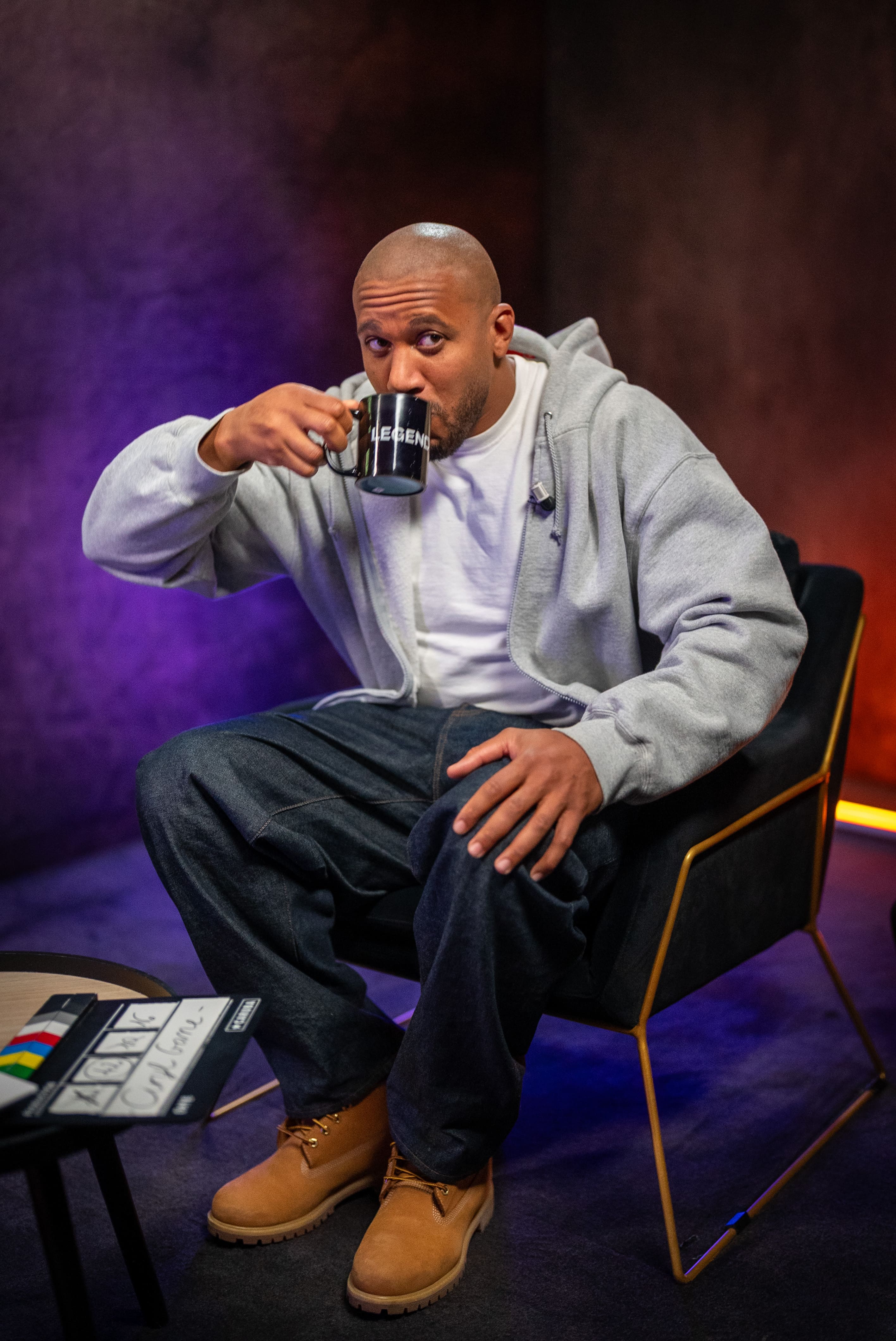 Photographie professionnelle du combattant de MMA français, Cyril Gane, en pleine interview pour Legend. Cyril Gane est assis sur un fauteuil moderne, buvant dans une tasse noire 'LEGEND'. Il est habillé de façon décontractée (sweat à capuche gris, t-shirt blanc, jean sombre, bottes camel). La lumière est tamisée avec des tons d'ambiance violets et rouges, typiques d'un studio d'interview. Cette photo de sport/lifestyle, optimisée par retouche, illustre l'expertise de Nux-Studio en production photo, direction artistique et création de contenu de marque pour des personnalités du monde du sport.