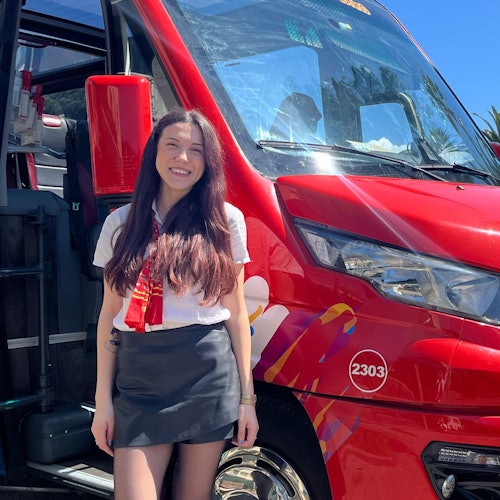 Smiling woman with long hair stands next to a red bus with colorful decals and the number 2303, under a clear blue sky.