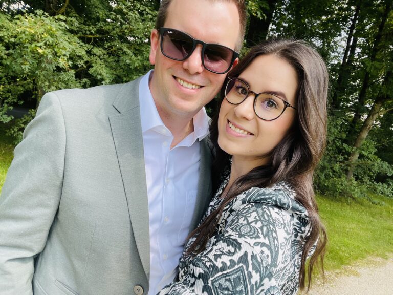 A smiling man with curly hair is sitting at a desk, looking at the camera, with a laptop and a lamp in the background.