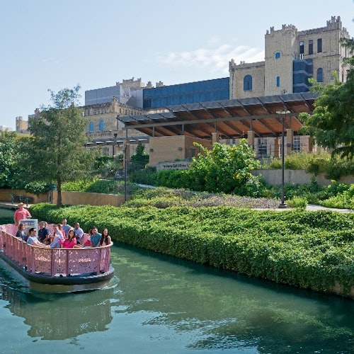 People ride a pink boat on a canal near lush greenery, with a large historic building in the background.