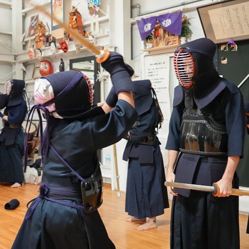 Twee personen in kendo-uitrusting oefenen met bamboezwaarden in een dojo, met extra beoefenaars op de achtergrond.