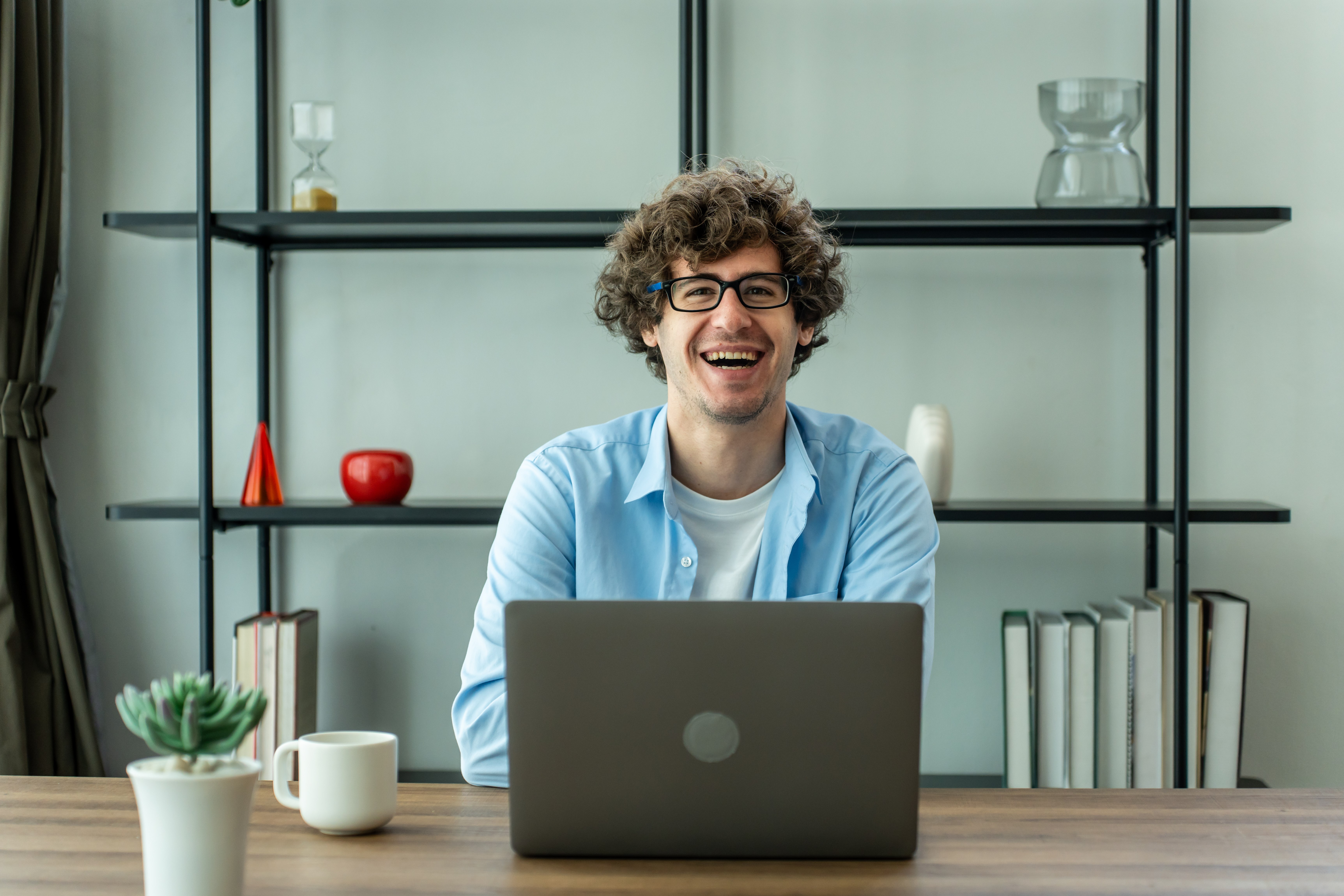 Ein junger, lächelnder Mann mit lockigen Haaren und Brille sitzt an seinem Schreibtisch, hinter dem sich ein Regal mit Büchern und Dekoration befindet. Er trägt ein hellblaues Hemd über einem weißen T-Shirt und blickt direkt in die Kamera, während ein Laptop vor ihm steht.