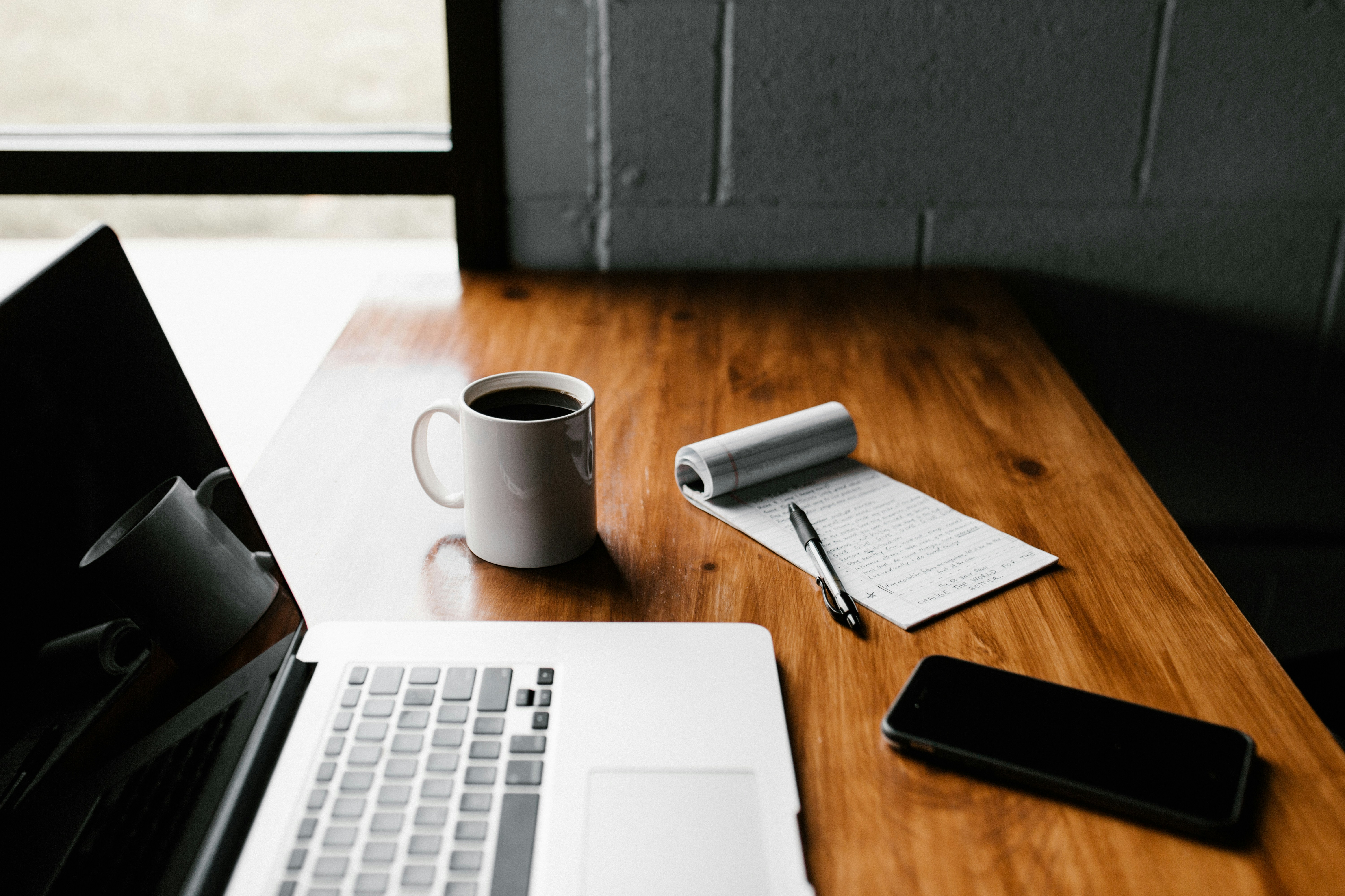 On a wooden table sits an open silver laptop with a blank screen. Behind it is a white mug of coffee and a pad and pen. A blank screen phone sits in front of the lapton.