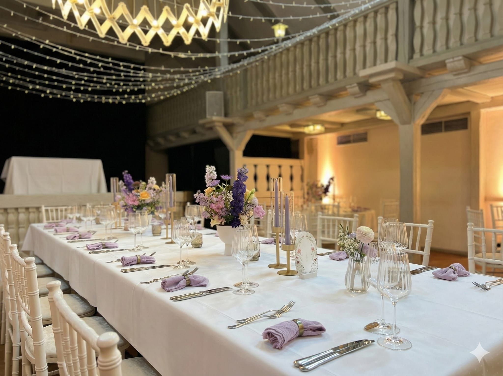 Lange Hochzeitstafel in einer hell erleuchteten Scheune mit dezenter Floristik und stimmungsvollen Lichterketten am Gebälk