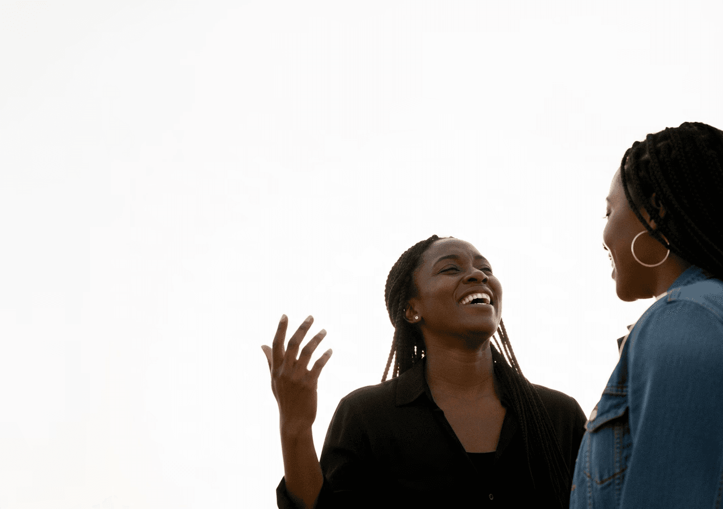 Two people with long braided hair are joyfully talking and gesturing against a clear sky backdrop.