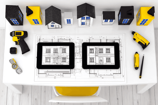 A white desk with house models, tools, and two tablets displaying architectural plans. The scene conveys a sense of design and creativity.