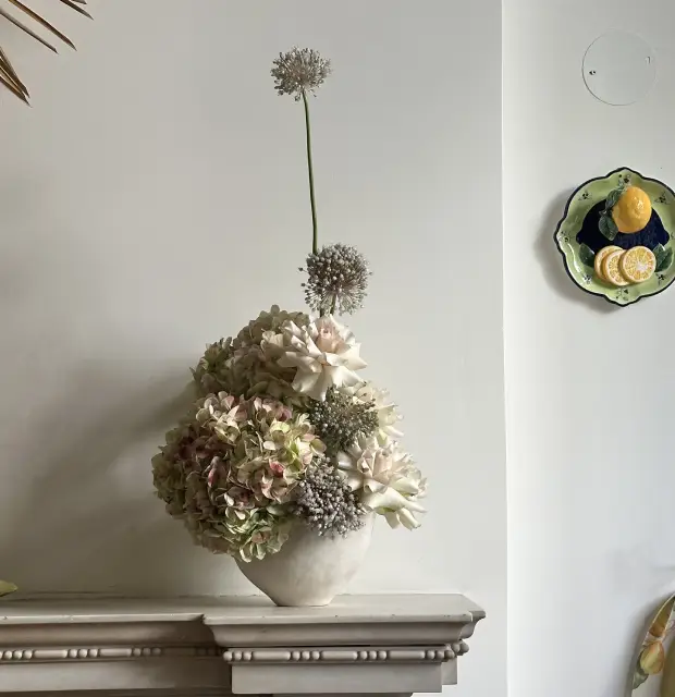Arreglo floral en tonos neutros con hortensias, dalias blancas y flores esféricas de allium en un jarrón de cerámica clara, sobre una repisa blanca frente a una pared minimalista.