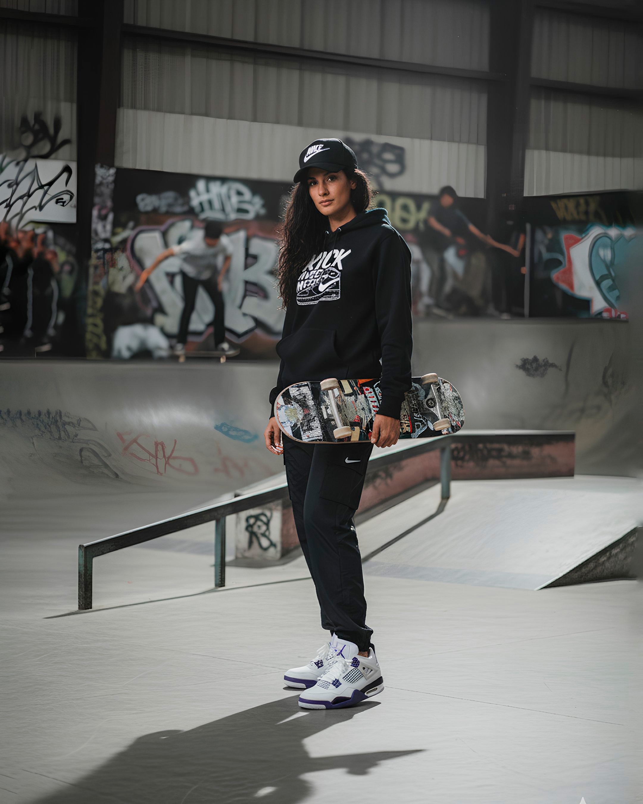 Portrait de mode lifestyle d'une jeune femme posant debout dans un skatepark urbain couvert. Elle porte une casquette noire Nike, un sweat à capuche noir avec un imprimé graphique blanc et un pantalon de survêtement noir, complétés par des sneakers blanches et violettes de type Jordan 4. Elle tient une planche de skateboard décorée de graffitis à hauteur de taille. L'arrière-plan montre des rampes de skate et des murs couverts de graffitis colorés, avec des skateurs flous en action, créant une ambiance de culture urbaine authentique.