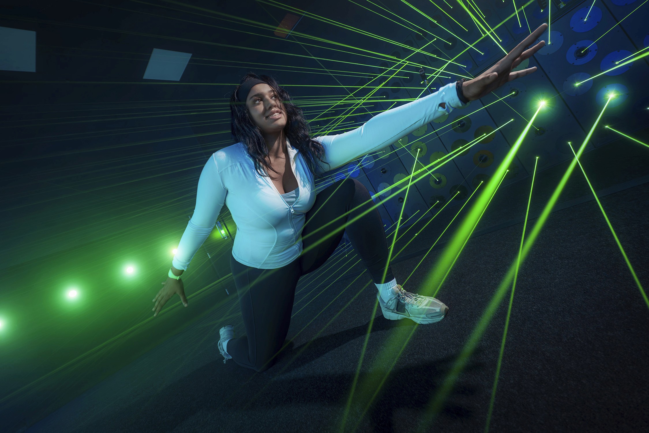 Woman kneeling, reaching out amidst a web of green laser beams in a dark room.