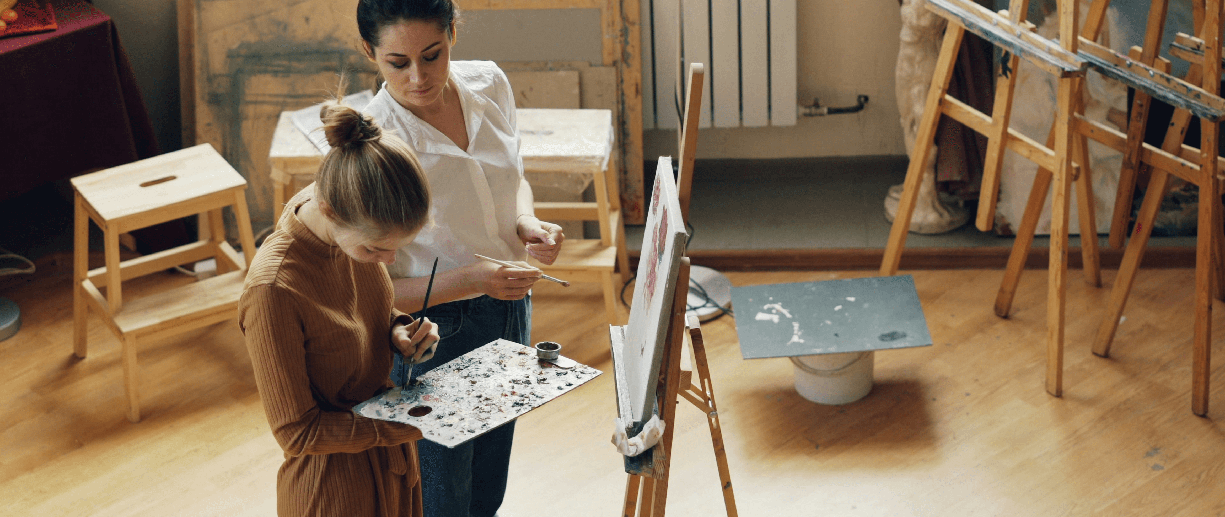 A person observes a canvas while another stands nearby, both in a sunlit creative space filled with art supplies.
