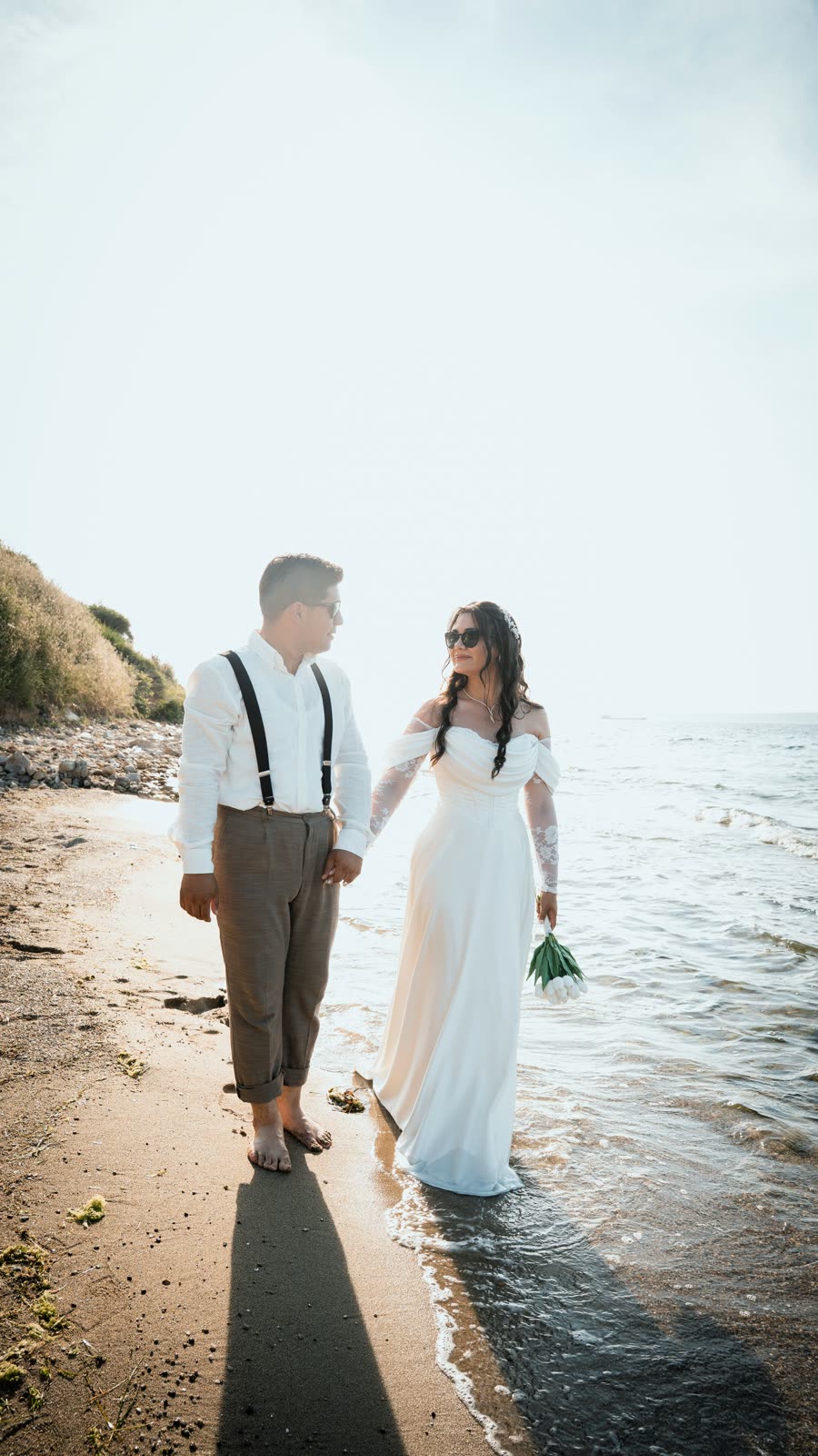 Couple walking in shallow water by a hillside with the bride holding a bouquet
