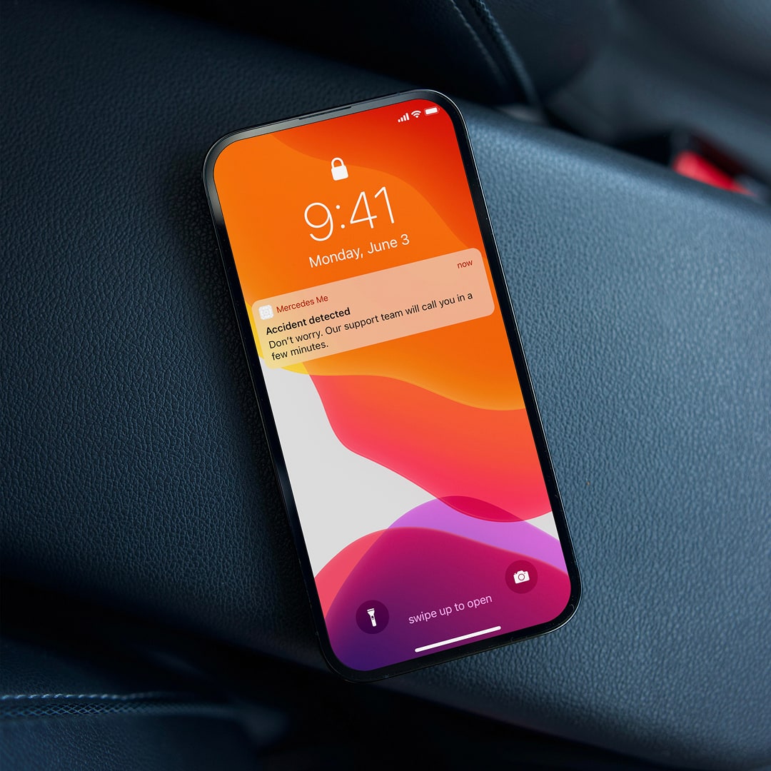 An iPhone resting on dark leather car upholstery showing a lock screen push notification from the Mercedes Me app reading "Accident detected — Don't worry. Our support team will call you in a few minutes."
