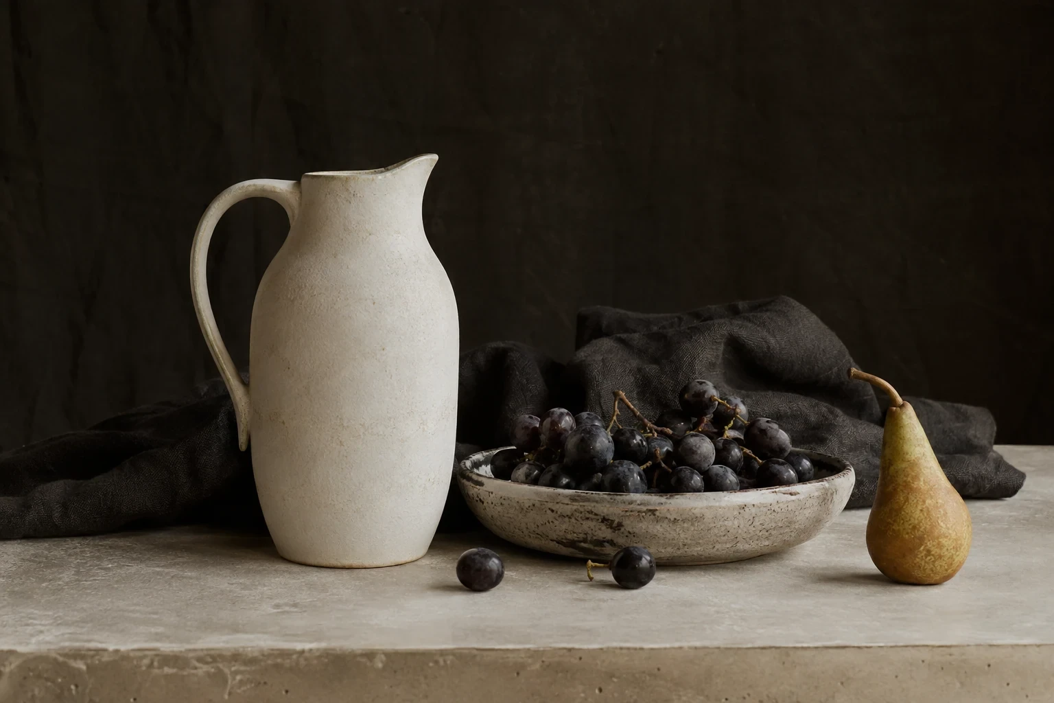 Ivory pottery pitcher with dark grapes in a low ceramic dish and a pear, dark cloth backdrop, pale tabletop