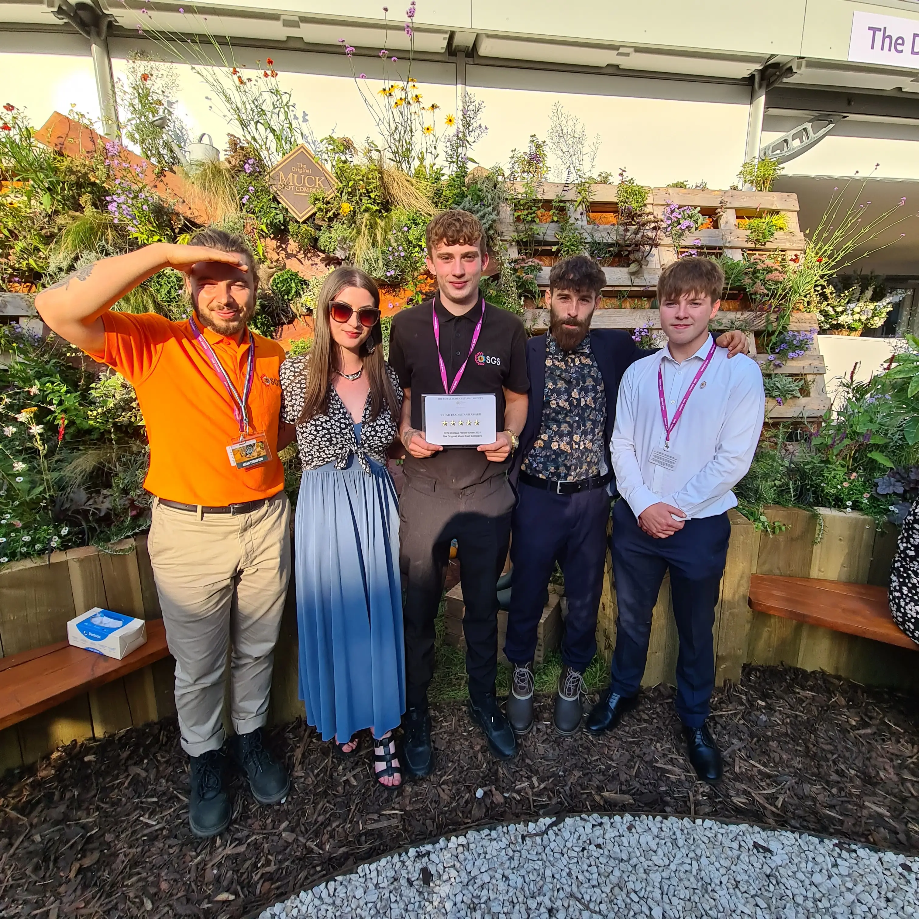 A group of four people stands outdoors, smiling and holding a certificate amid greenery.