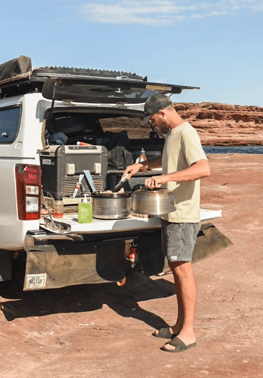 Parrilla COBB Supreme sobre una mesa lista para cocinar en una playa,  detrás se ven una sillas y una camioneta sobre la arena