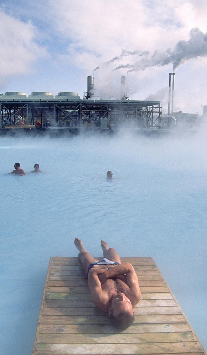 Man relaxes on a wooden platform by a steaming geothermal pool, with industrial plant and smoke stacks in the background, under a cloudy sky.