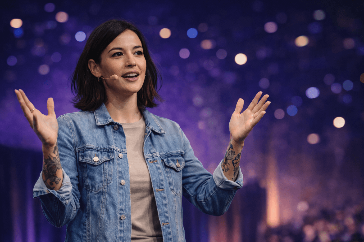 Woman in denim jacket speaking on stage with her hands raised.