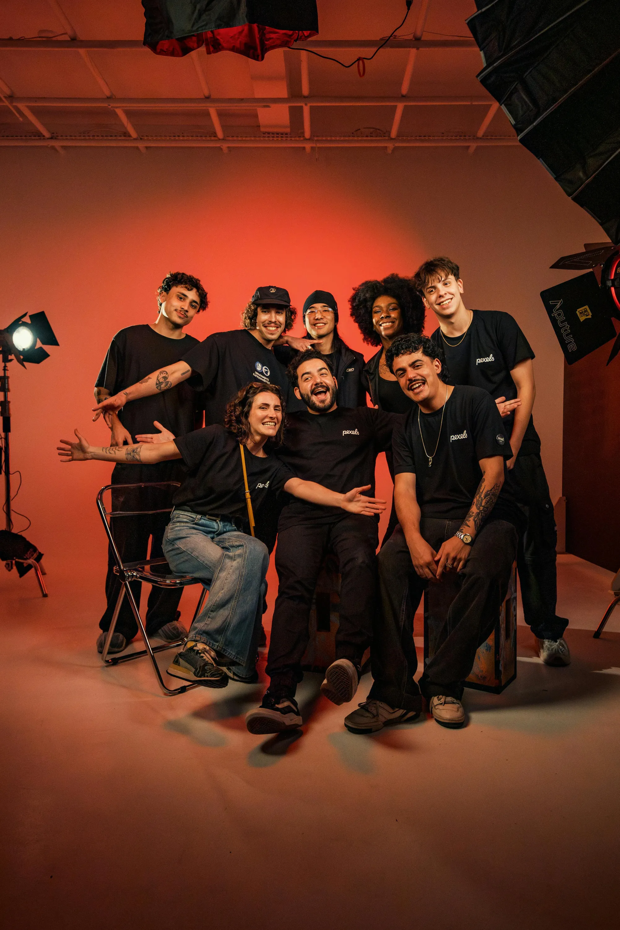 Eight people in black Pexels shirts posing happily in a photo studio with red lighting and professional equipment.