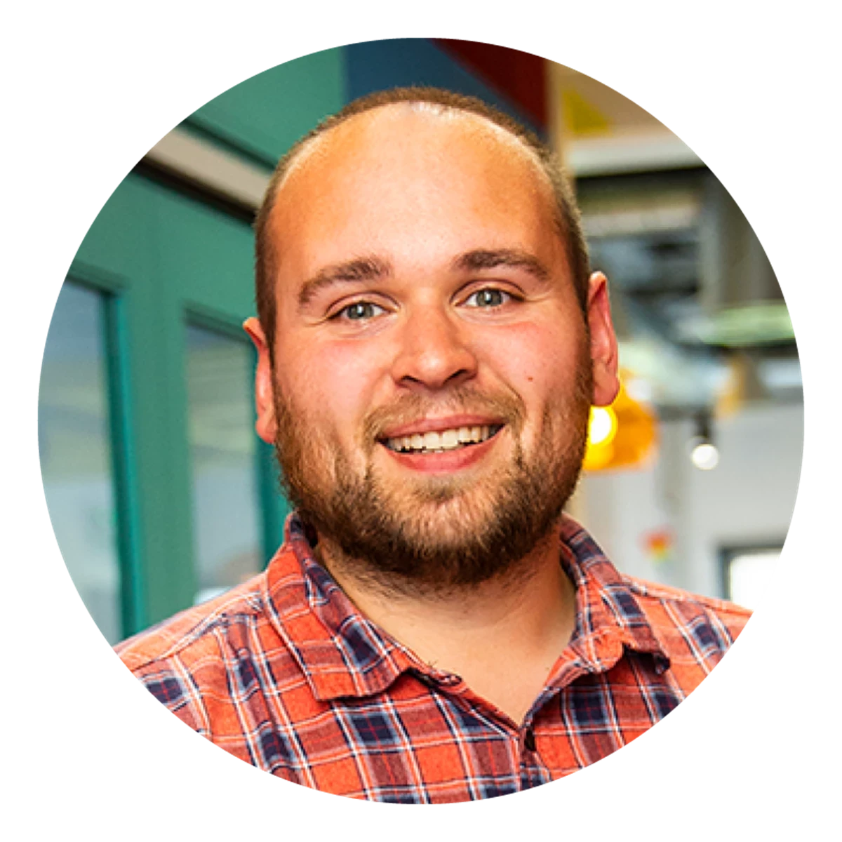 A circular portrait shows a person wearing a red‑and‑blue checked shirt in a bright office environment. Coloured walls and hanging lights appear in the background.