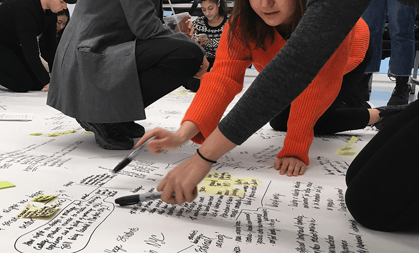 A diverse group of researchers, designers, strategists, and community members kneel on the floor co-creating a large systems map covered in handwritten insights and post-its during a workshop.