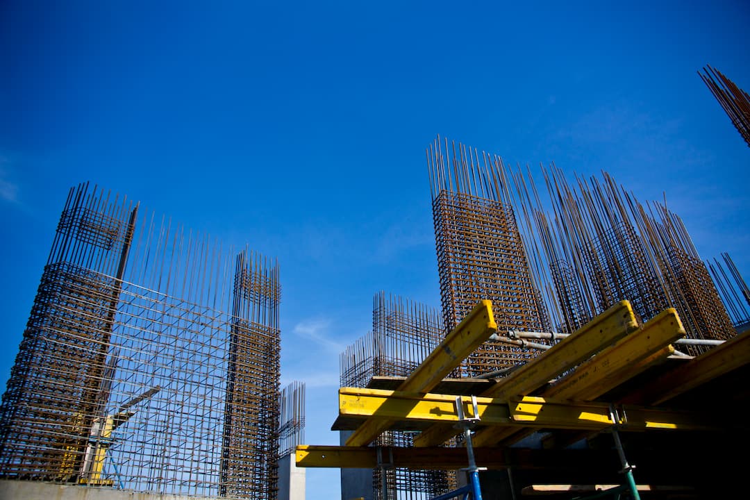 Steel rebar cages rise from a construction site foundation, with yellow formwork beams and scaffolding in the foreground against a clear blue sky.