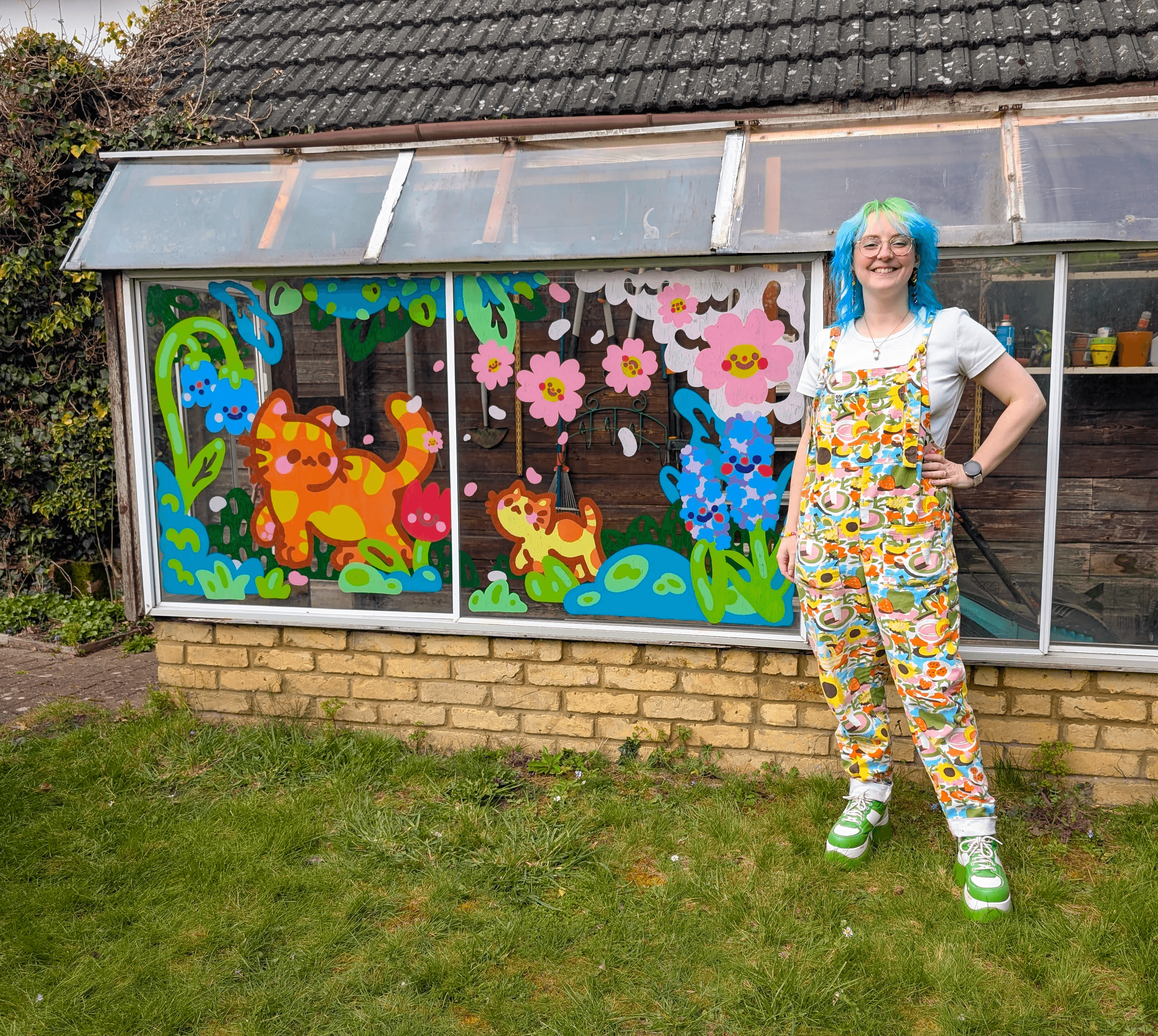 A low greenhouse window decorated with a large, colourful illustration of a ginger cat, looking back at a small ginger kitten. They are walking through a spring scene of smiling flowers and greenery. The artist, a white woman with blue and green hair, stands next to the work.