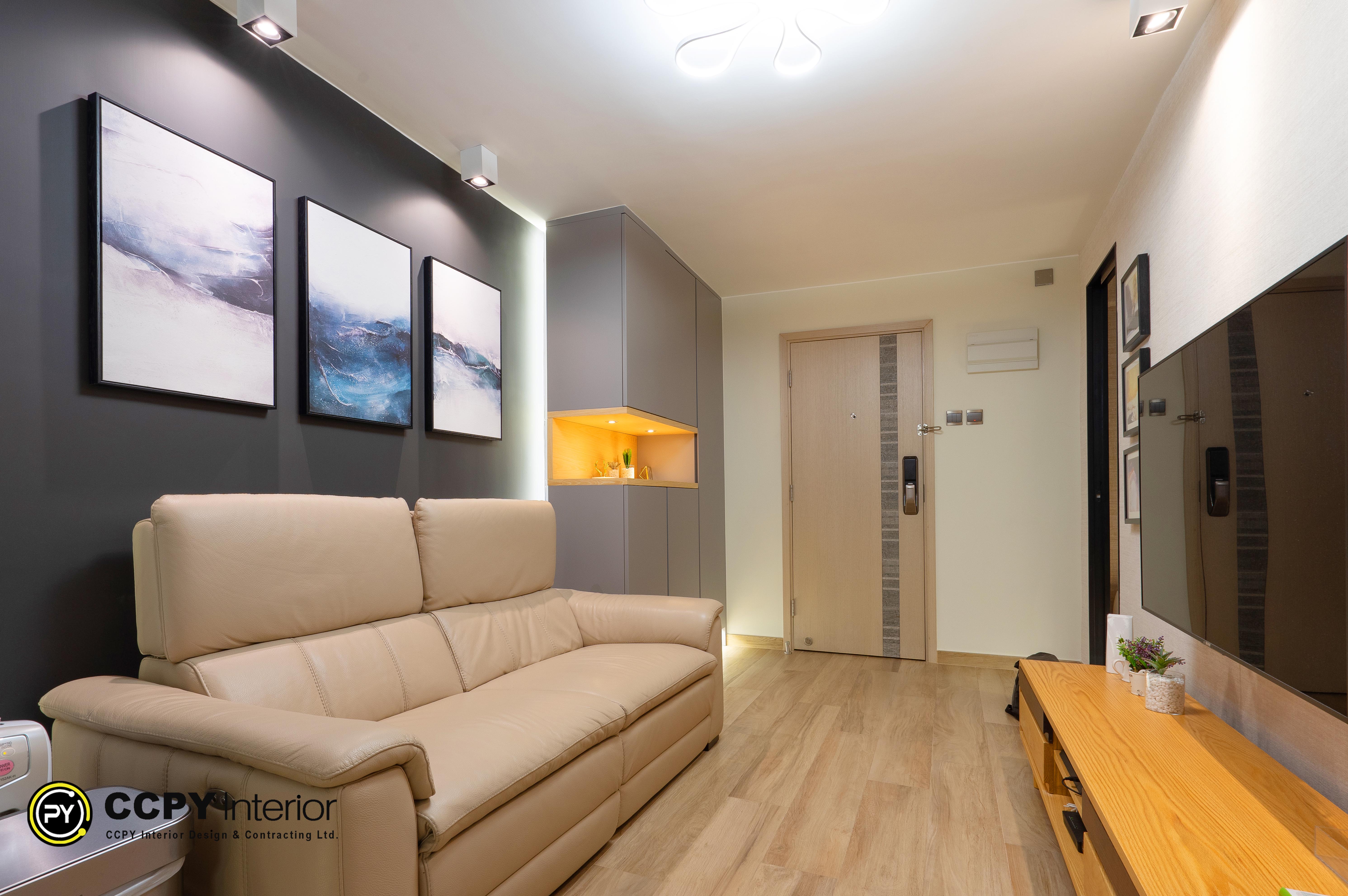 This is a modern living room interior featuring a beige leather sofa against a charcoal grey feature wall adorned with three abstract landscape artworks.Lighting: The space utilizes hidden LED strip lighting to highlight the feature wall and a warm spotlight within a recessed cabinet nook.Furniture: A wooden TV console sits opposite the sofa, complemented by a light wood-toned floor that matches the overall aesthetic.Color Palette: The design combines a neutral beige sofa with contrasting dark grey walls and light wood flooring to create a balanced, contemporary look