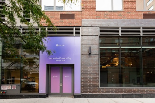 Street-level entrance of a modern brick building featuring a purple event banner that reads “Pyth – Innovative Finance Day, Mainnet 2024.” The glass doors and large adjacent windows reflect surrounding urban elements, while a tree branch extends partly into the frame from the left.