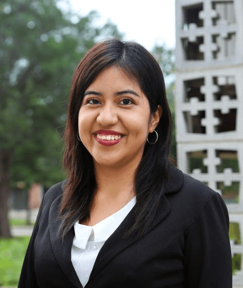 Smiling woman with dark hair wearing a black blazer and white blouse. She stands outdoors with geometric sculpture and trees in the background
