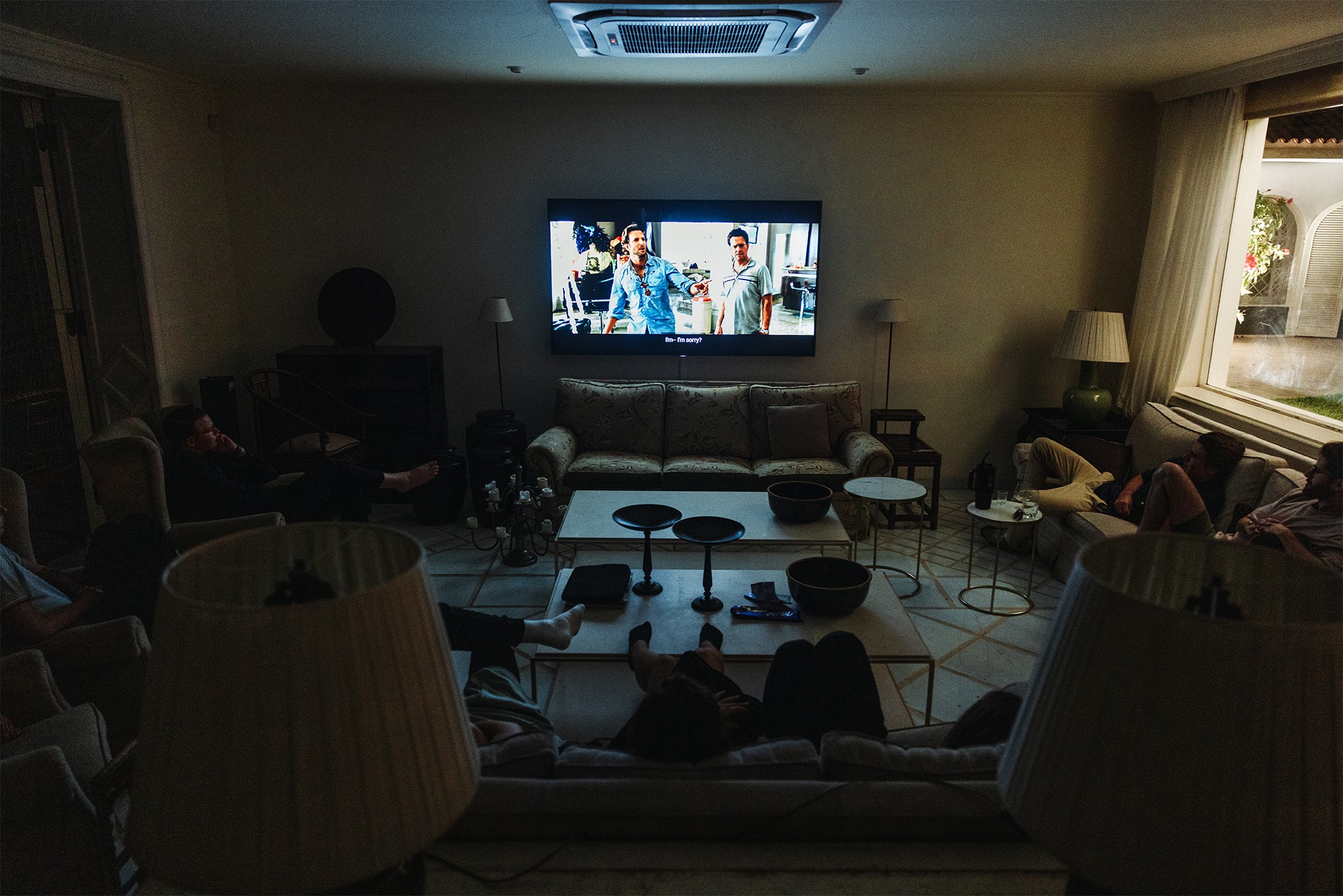 A dimly lit living room with people watching a movie on a wall-mounted television