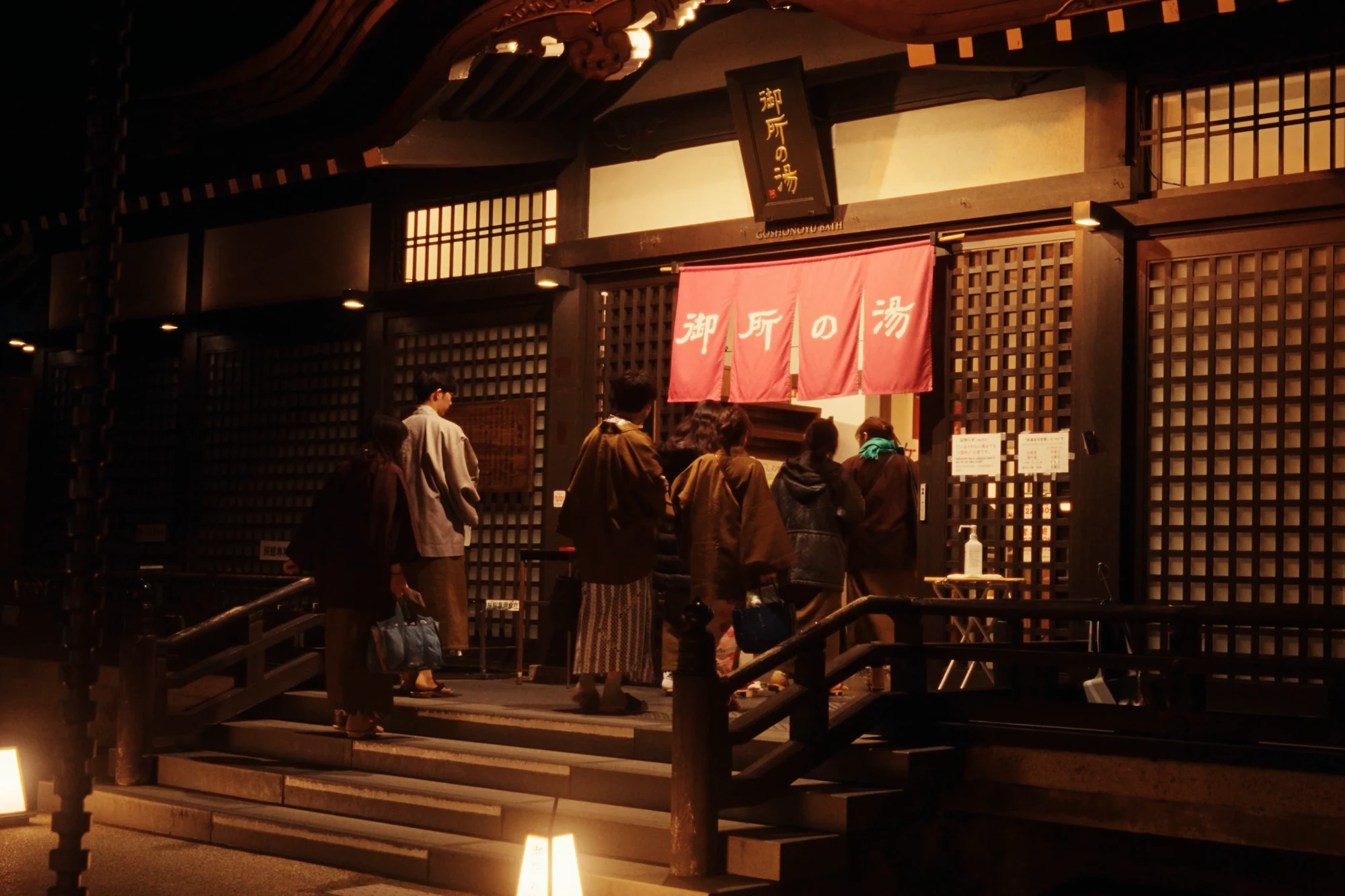 Bathers in yukata entering Goshono-yu bathhouse at night under lanterns.