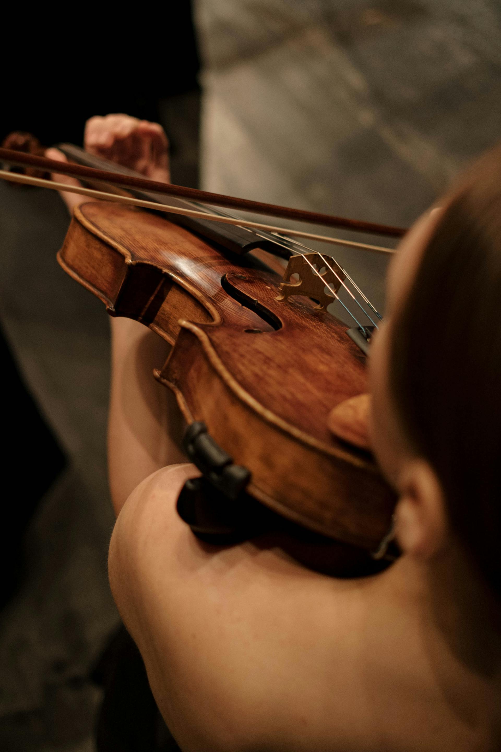 An over-the-shoulder perspective of a violinist focusing on the strings and bow movement across the body of the wooden instrument.